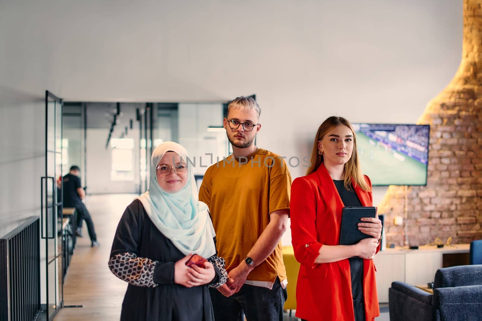 A group of young business colleagues, including a woman in a hijab, stands united in the modern corridor of a spacious startup coworking center