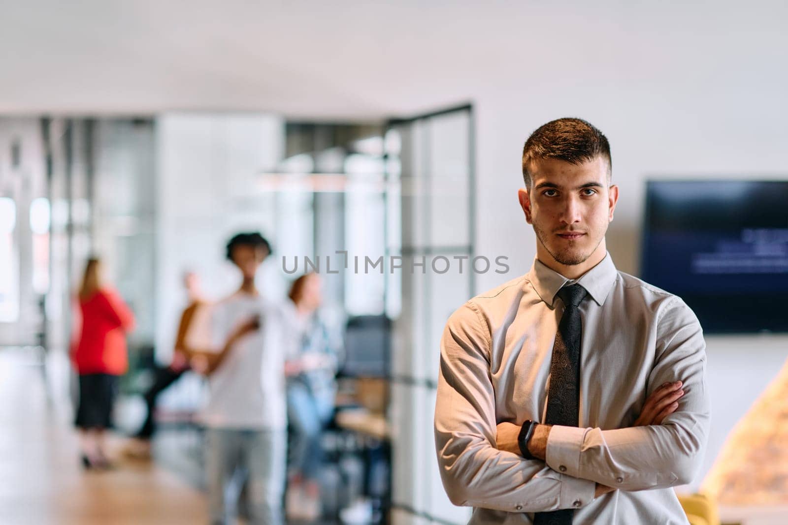 A young business leader stands with crossed arms in a modern office hallway, radiating confidence and a sense of purpose, embodying a dynamic and inspirational presence.
