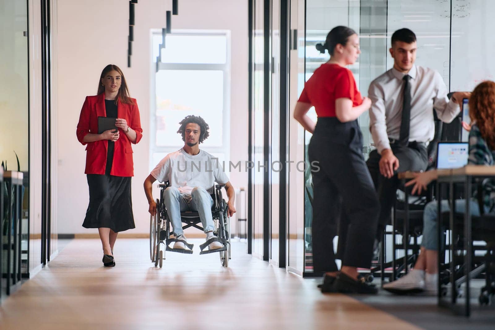 A business leader with her colleague, an African-American businessman who is a disabled person, pass by their colleagues who work in modern offices.