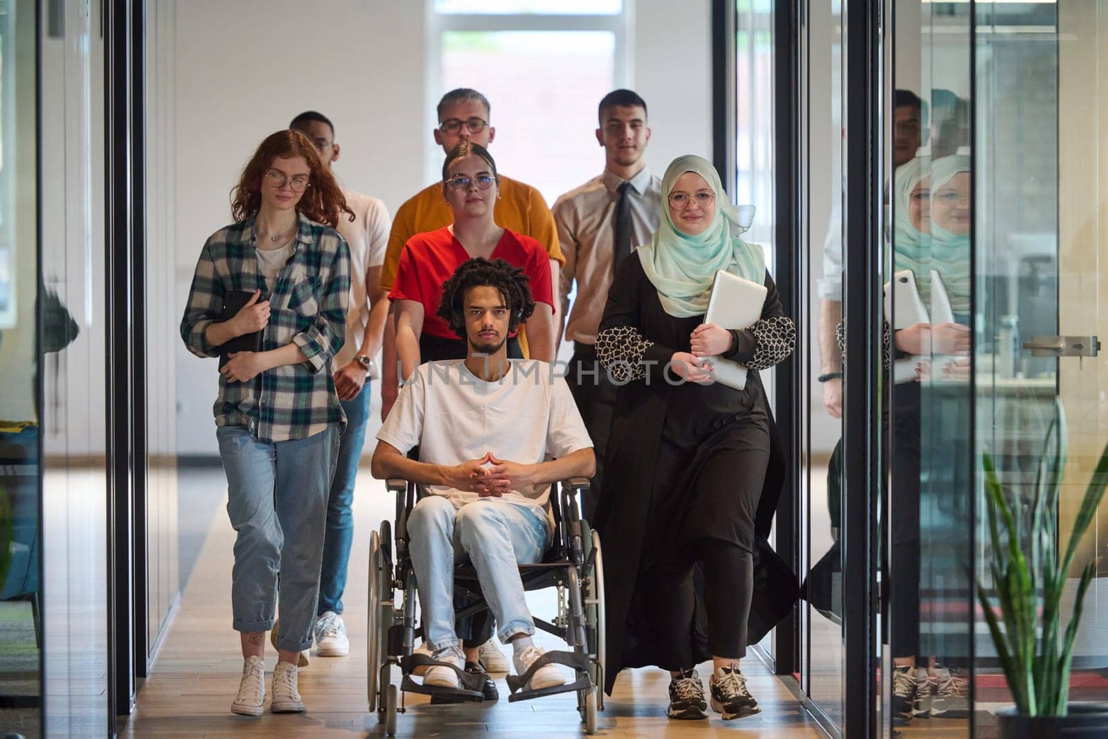 A diverse group of young business people congregates within a modern startup's glass-enclosed office, featuring inclusivity with a person in a wheelchair, an African American young man, and a hijab muslim woman . by dotshock
