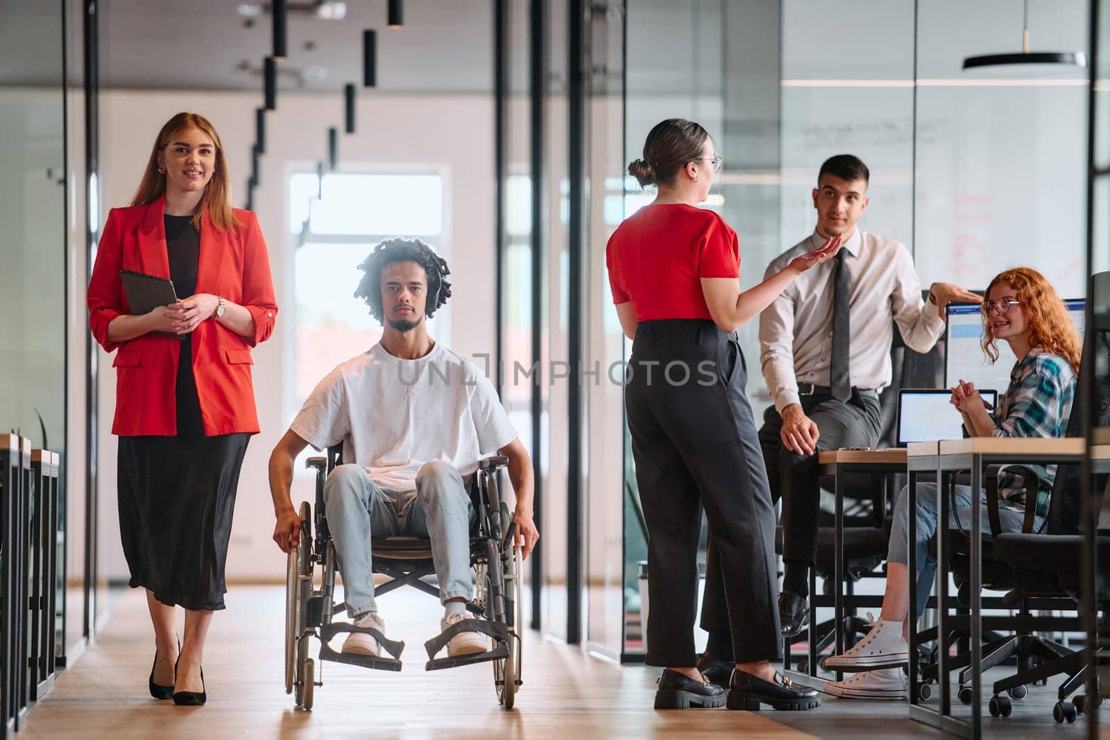 A business leader with her colleague, an African-American businessman who is a disabled person, pass by their colleagues who work in modern offices.