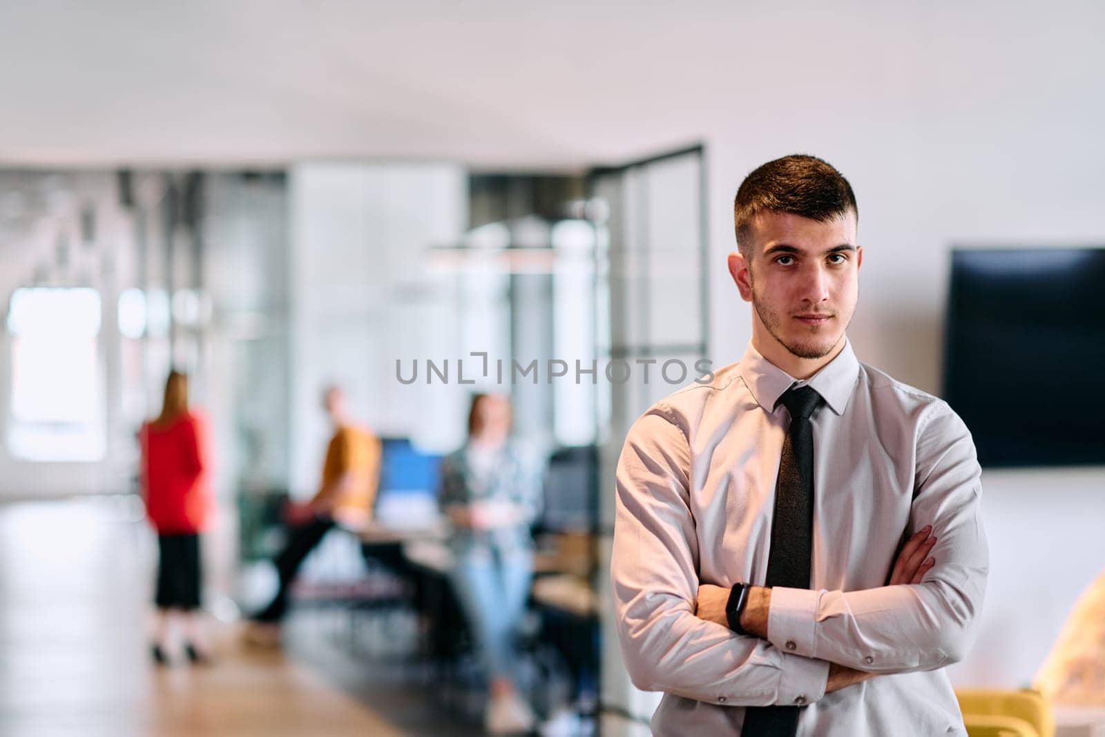 A young business leader stands with crossed arms in a modern office hallway, radiating confidence and a sense of purpose, embodying a dynamic and inspirational presence.