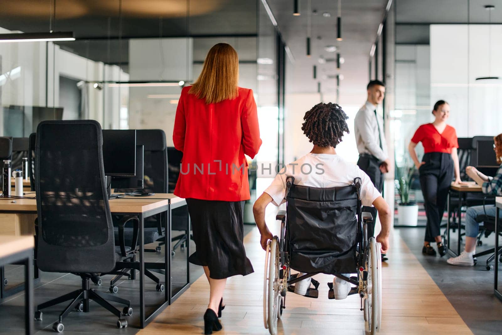 A business leader with her colleague, an African-American businessman who is a disabled person, pass by their colleagues who work in modern offices.