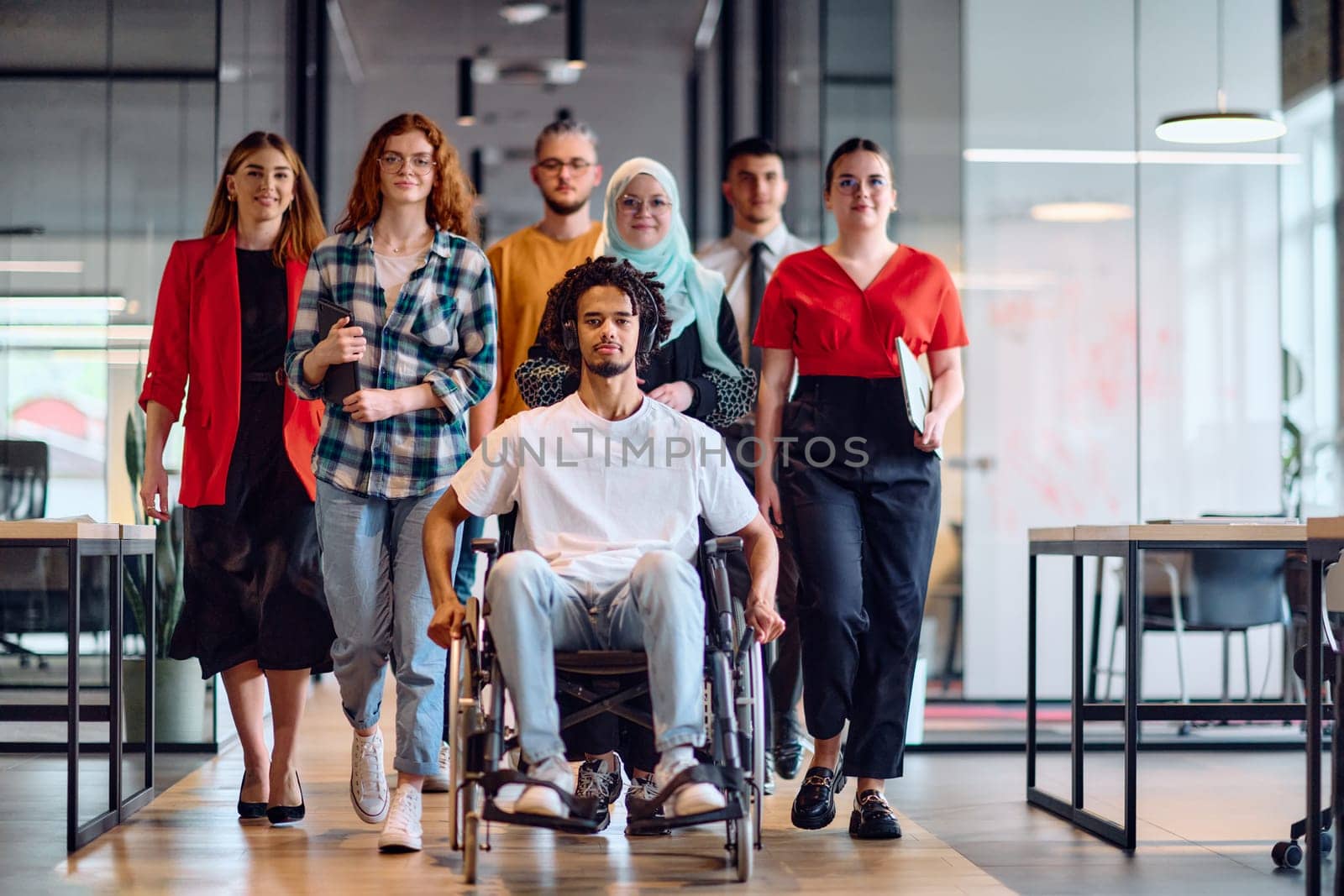 A diverse group of young business people walking a corridor in the glass-enclosed office of a modern startup, including a person in a wheelchair and a woman wearing a hijab by dotshock