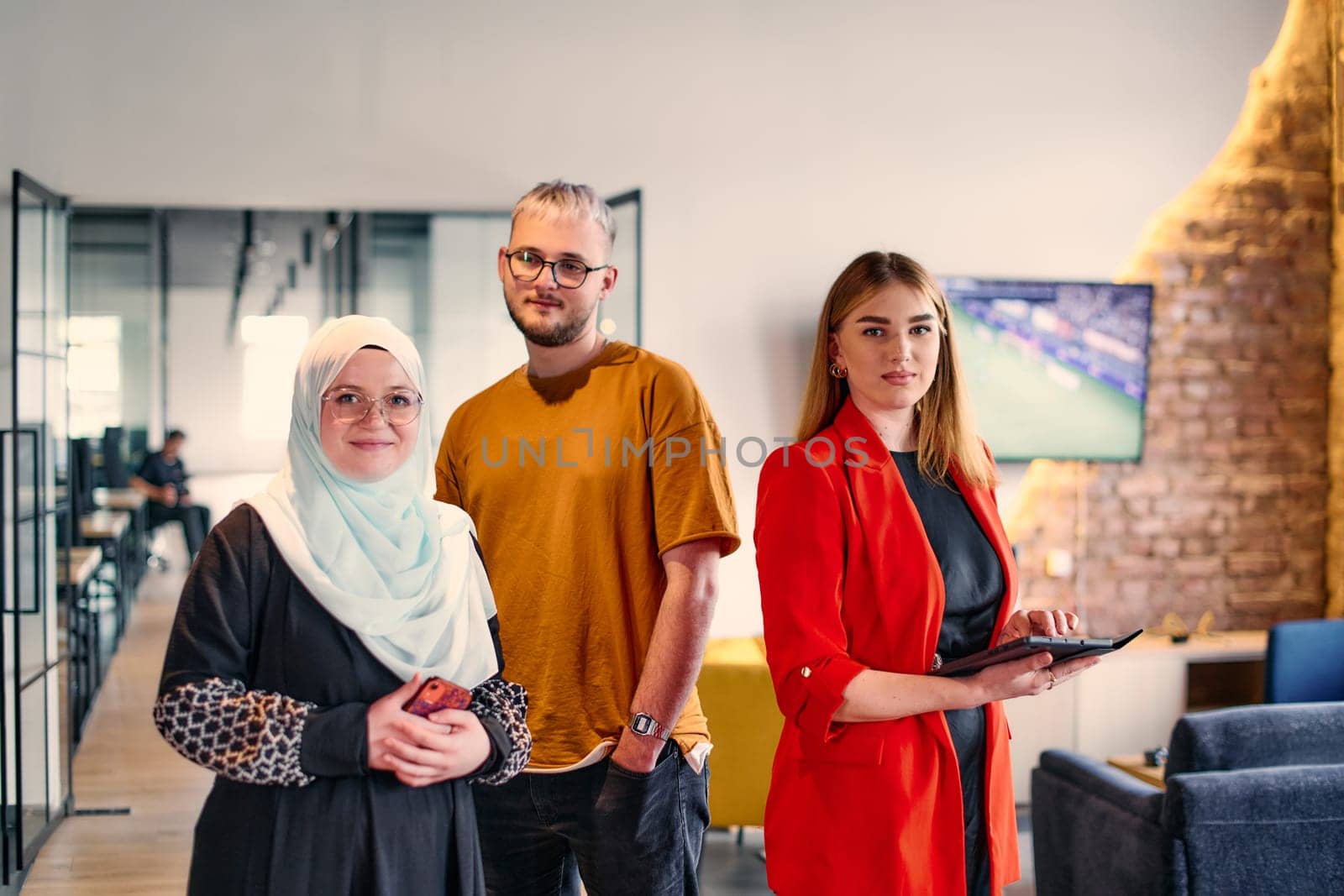 A group of young business colleagues, including a woman in a hijab, stands united in the modern corridor of a spacious startup coworking center
