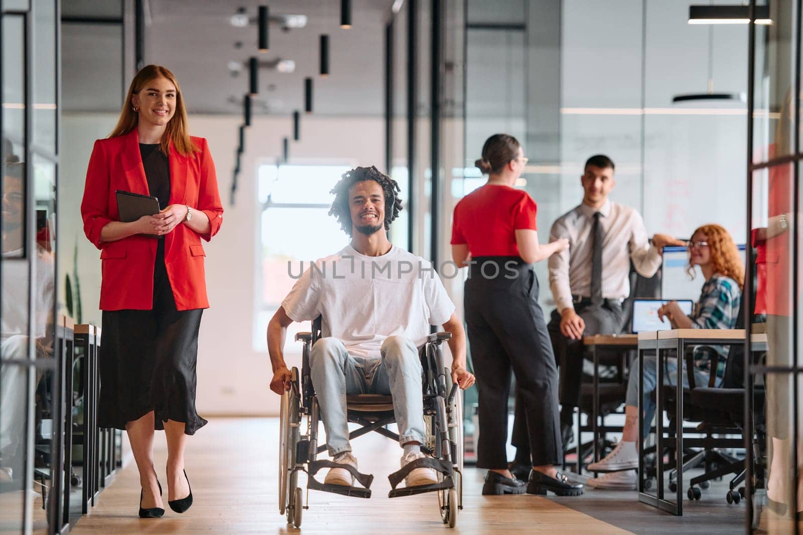 A business leader with her colleague, an African-American businessman who is a disabled person, pass by their colleagues who work in modern offices.