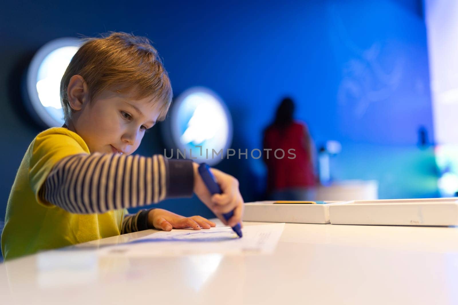 A boy is drawing on a piece of paper with a blue marker. He is wearing a yellow shirt