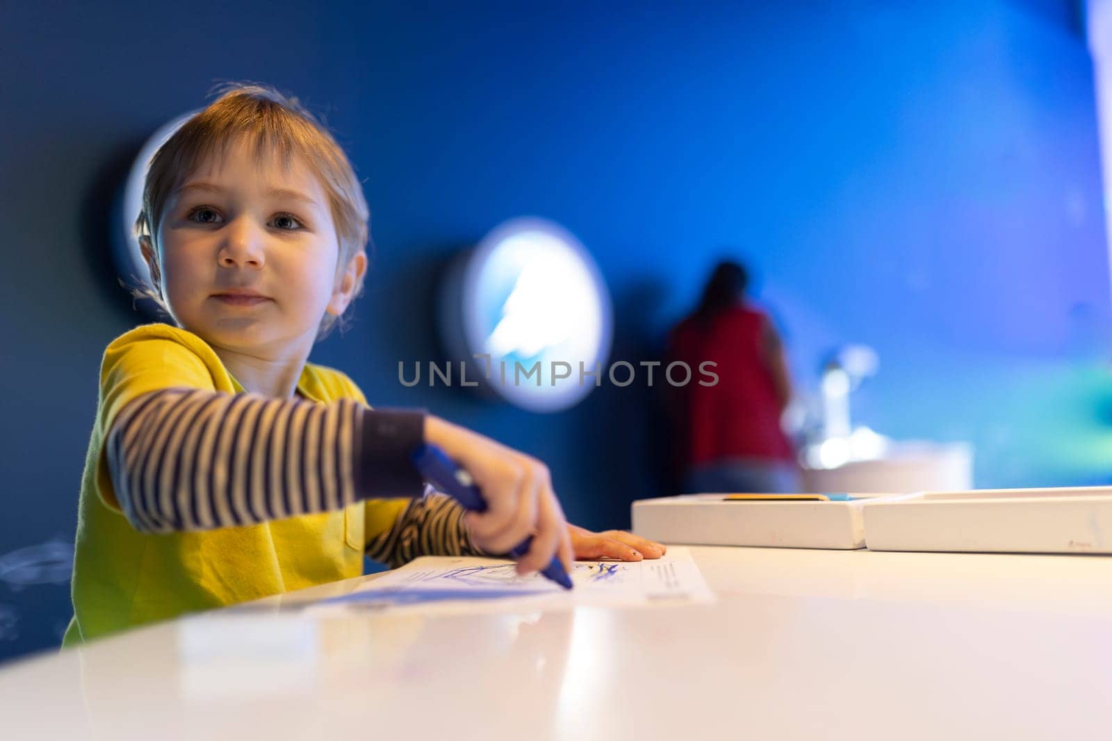 A young boy is sitting at a table with a blue marker in his hand. He is looking at a piece of paper and seems to be drawing or writing something. Concept of creativity and focus