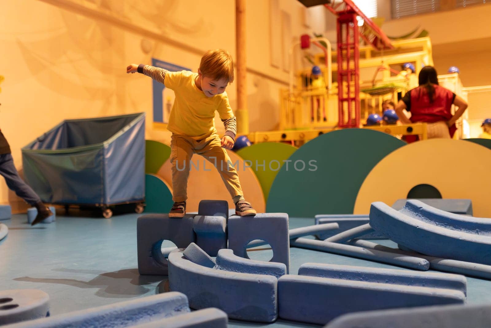 A young boy is playing with blocks in a room. The blocks are blue and the boy is standing on them. There are other people in the room, but they are not the main focus of the image