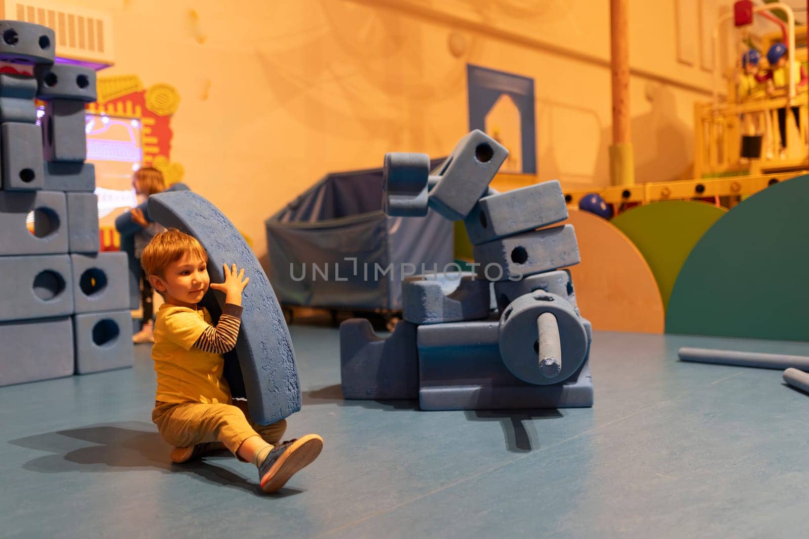 A young boy is playing with a blue toy block in a room with other toys. The boy is holding the block in his arms and he is enjoying himself. The room is filled with various toys, including a truck
