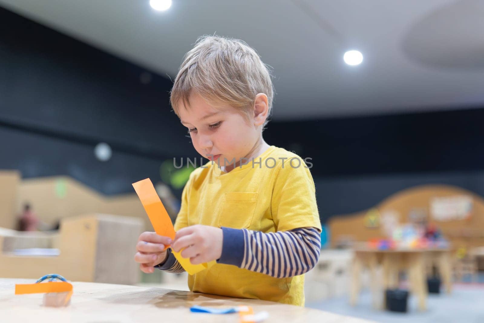 A young boy is sitting at a table with a piece of paper in his hand. He is focused on cutting the paper with scissors. The scene suggests a moment of concentration and creativity