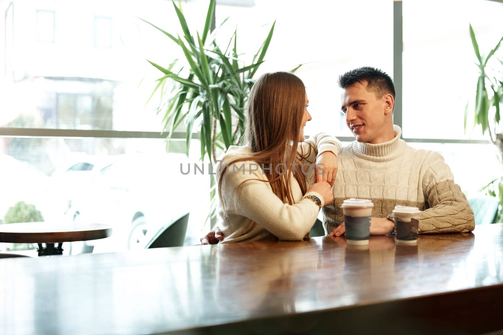 A couple sits close together at a wooden table in a brightly lit cafe, exchanging smiles over steaming cups of perhaps coffee or tea. They appear relaxed and engaged in a cheerful conversation, with natural light filtering through the space creating a warm and inviting atmosphere.
