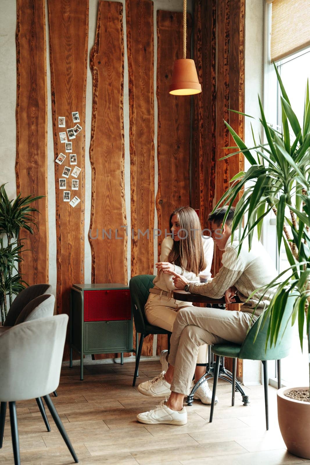 A smiling young couple sits closely together at a small table in a warmly lit cafe, sharing a moment over coffee. Surrounded by indoor plants and rustic decor, they seem engaged in a pleasant conversation, with a backdrop of draping curtains and a casual atmosphere.