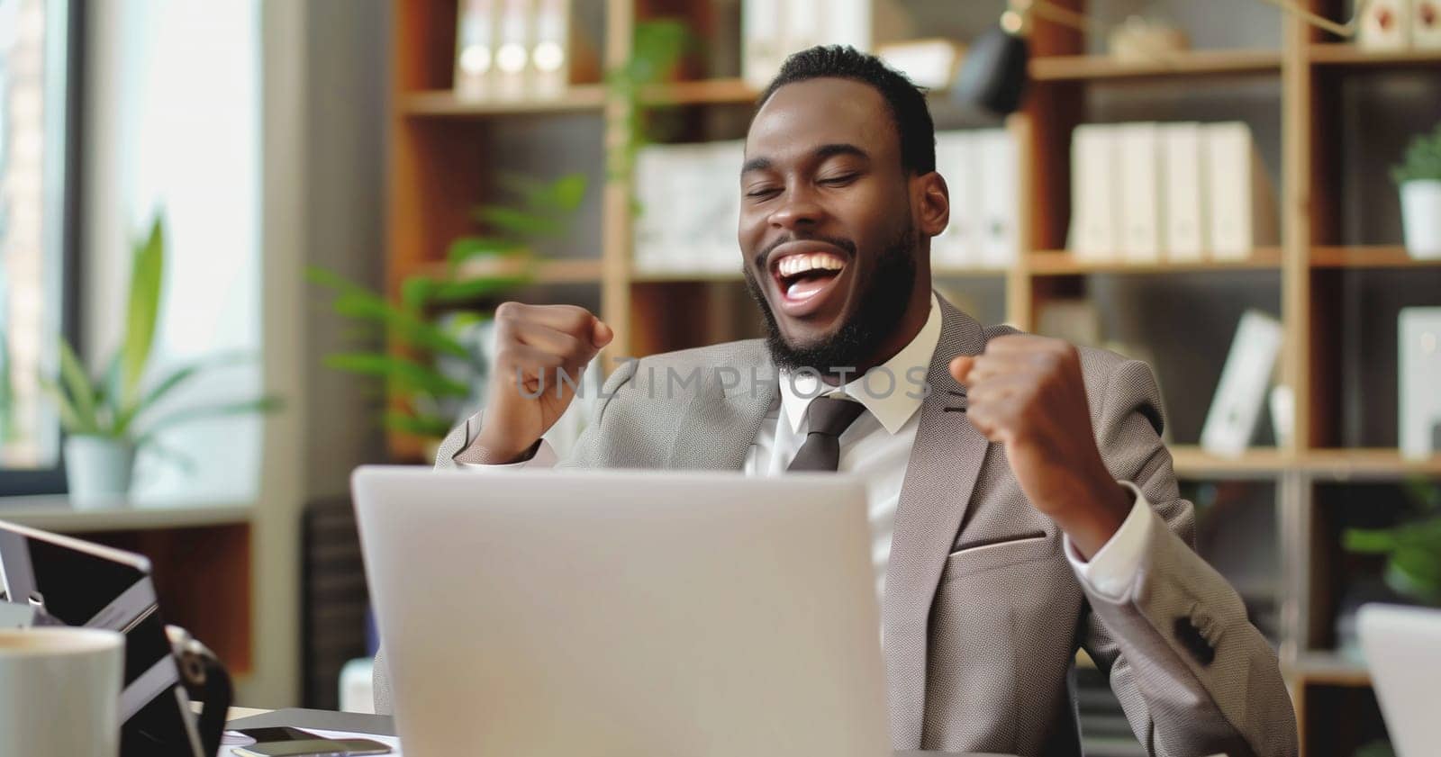 Happy overjoyed laughing african businessman celebrating success raising his hands up while working on laptop sitting at his desk in the office