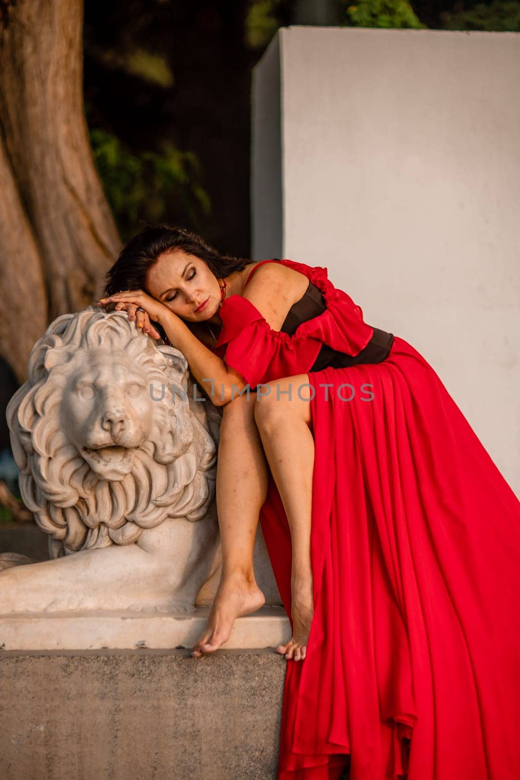 A woman in a red dress is sitting on a stone lion statue. The statue is white and has a lion's head. The woman is wearing a black belt and is looking at the camera. The scene has a calm