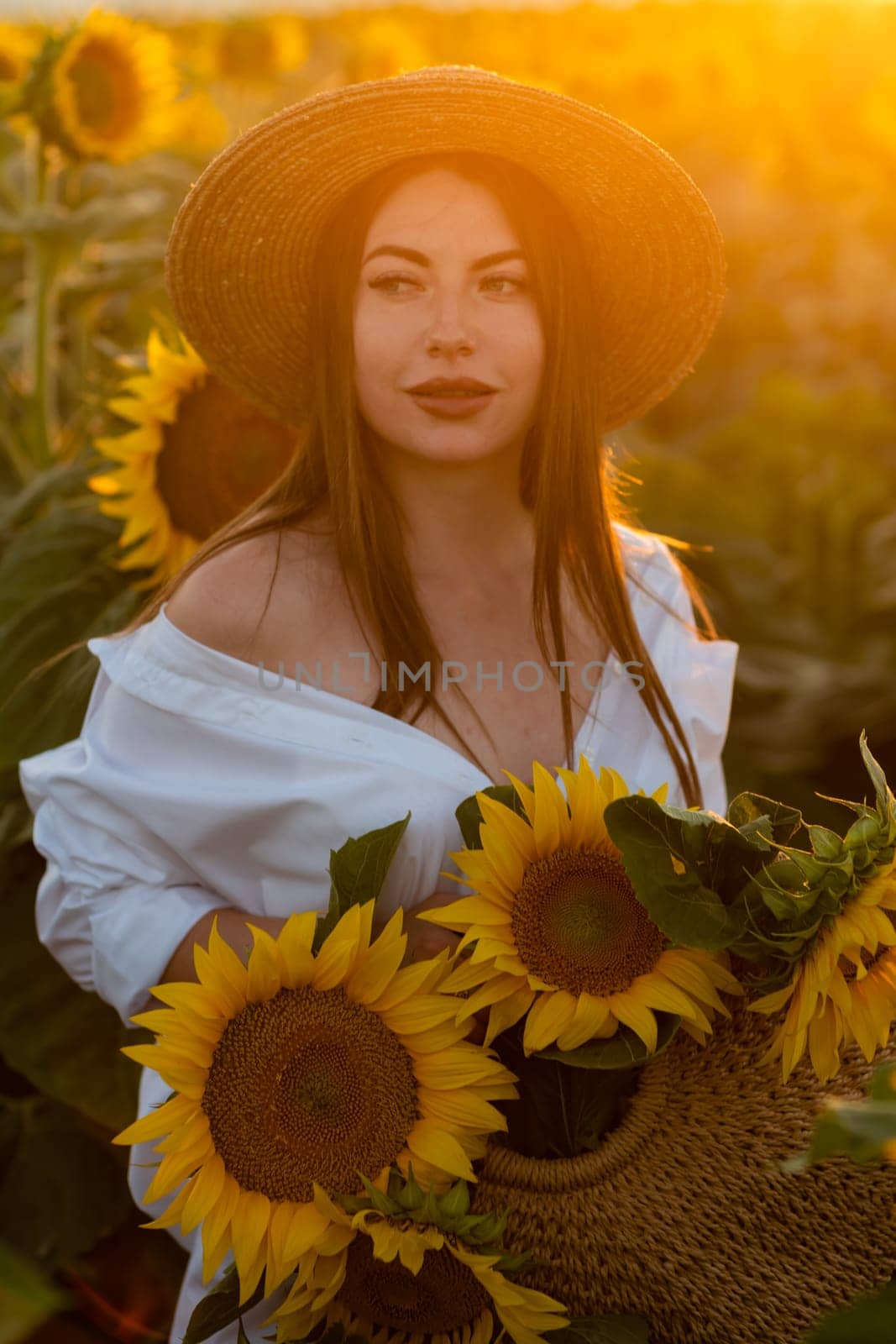 A girl in a hat on a beautiful field of sunflowers against the sky in the evening light of a summer sunset. Sunbeams through the flower field. Natural background. by Matiunina