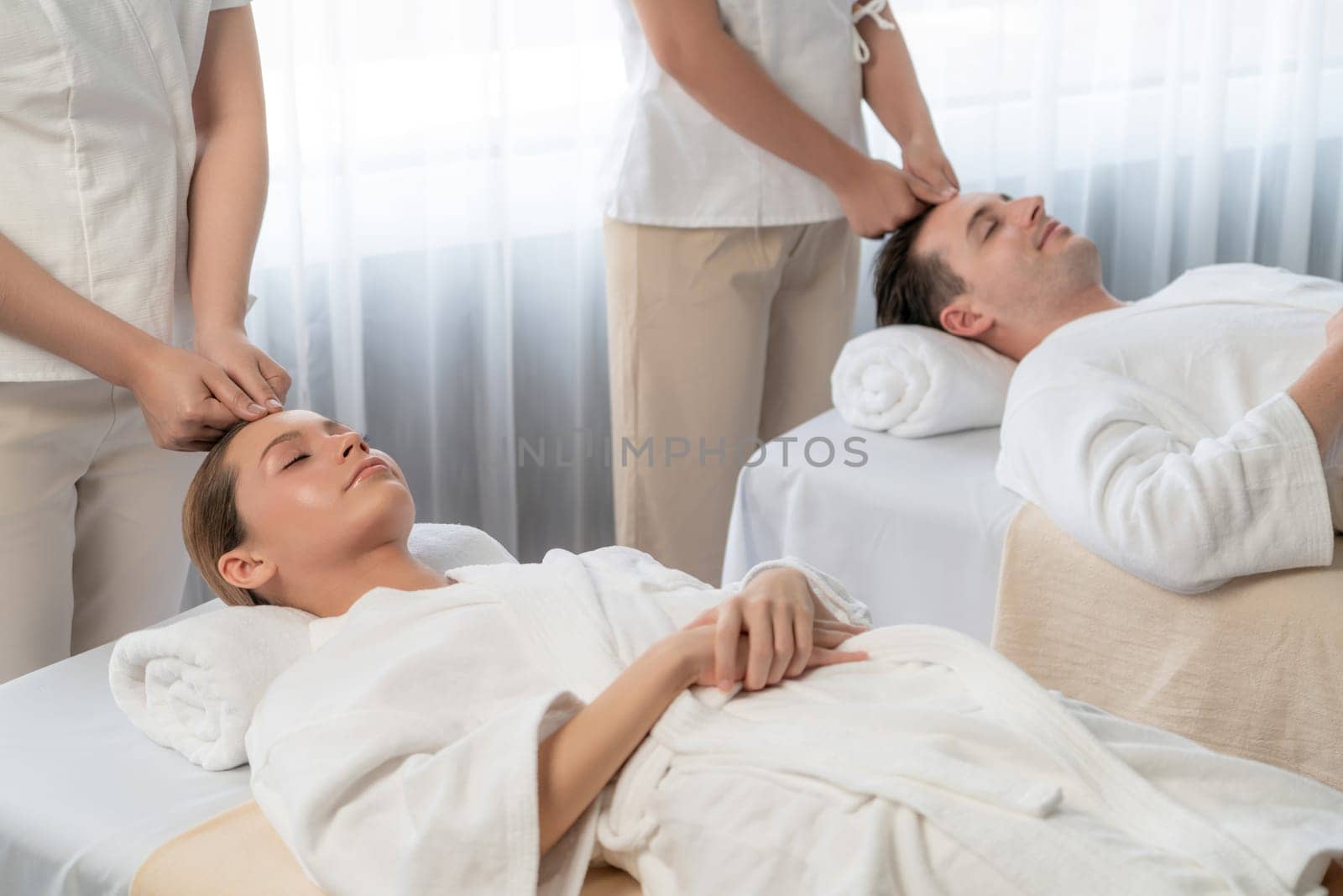 Caucasian couple enjoying relaxing anti-stress head massage and pampering facial beauty skin recreation leisure in dayspa modern light ambient at luxury resort or hotel spa salon. Quiescent