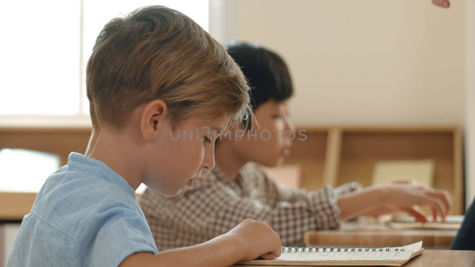 Side view of caucasian elementary student looking a note and answer question while sitting in front of blackboard. Young teacher explain classwork or test while children attend the class. Pedagogy.