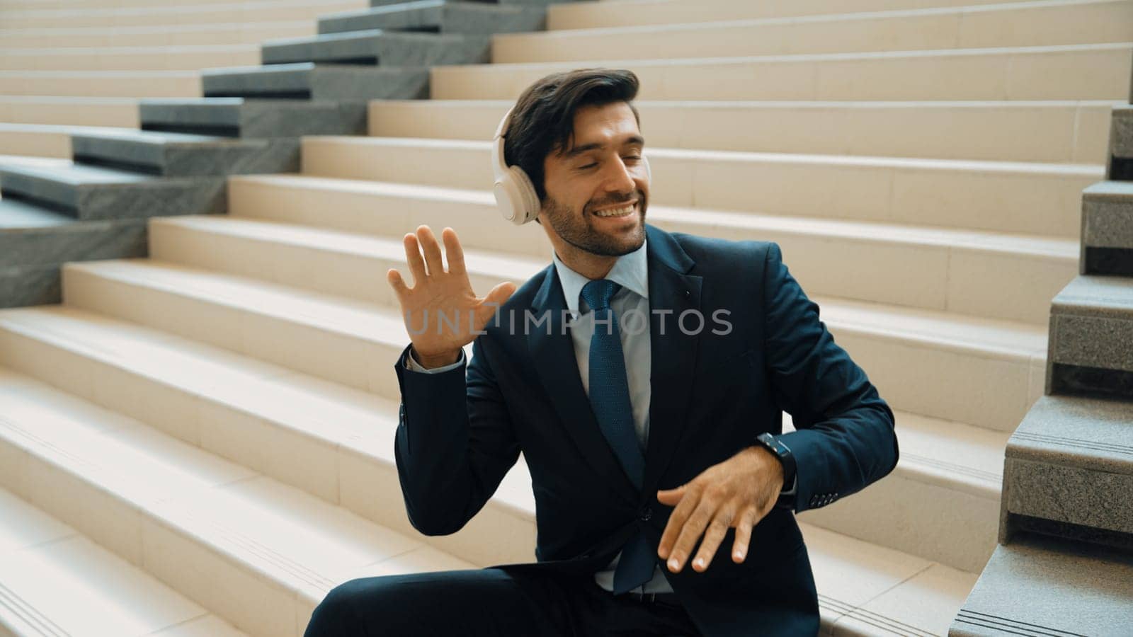 Project manager listening and enjoy music from headphone while sitting at stairs. Professional businessman wearing suit outfit while dancing and moving to music. Happy man listen funny song. Exultant.