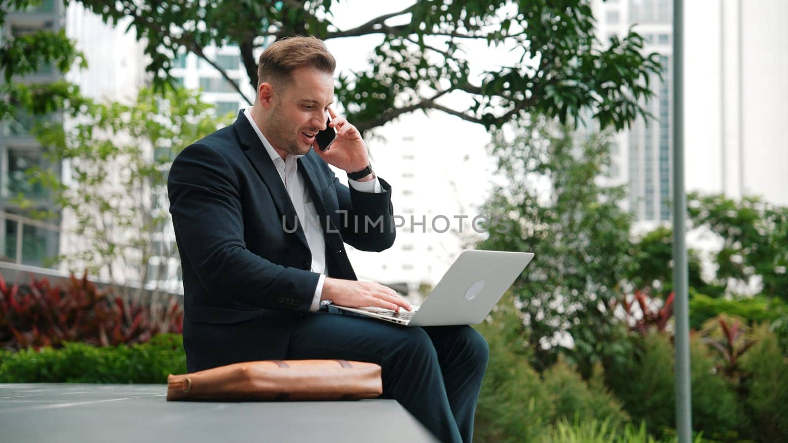 Caucasian businessman sitting at eco park while talking by using phone and working on laptop. Professional executive manager explain marketing idea from computer while calling to investor. Urbane