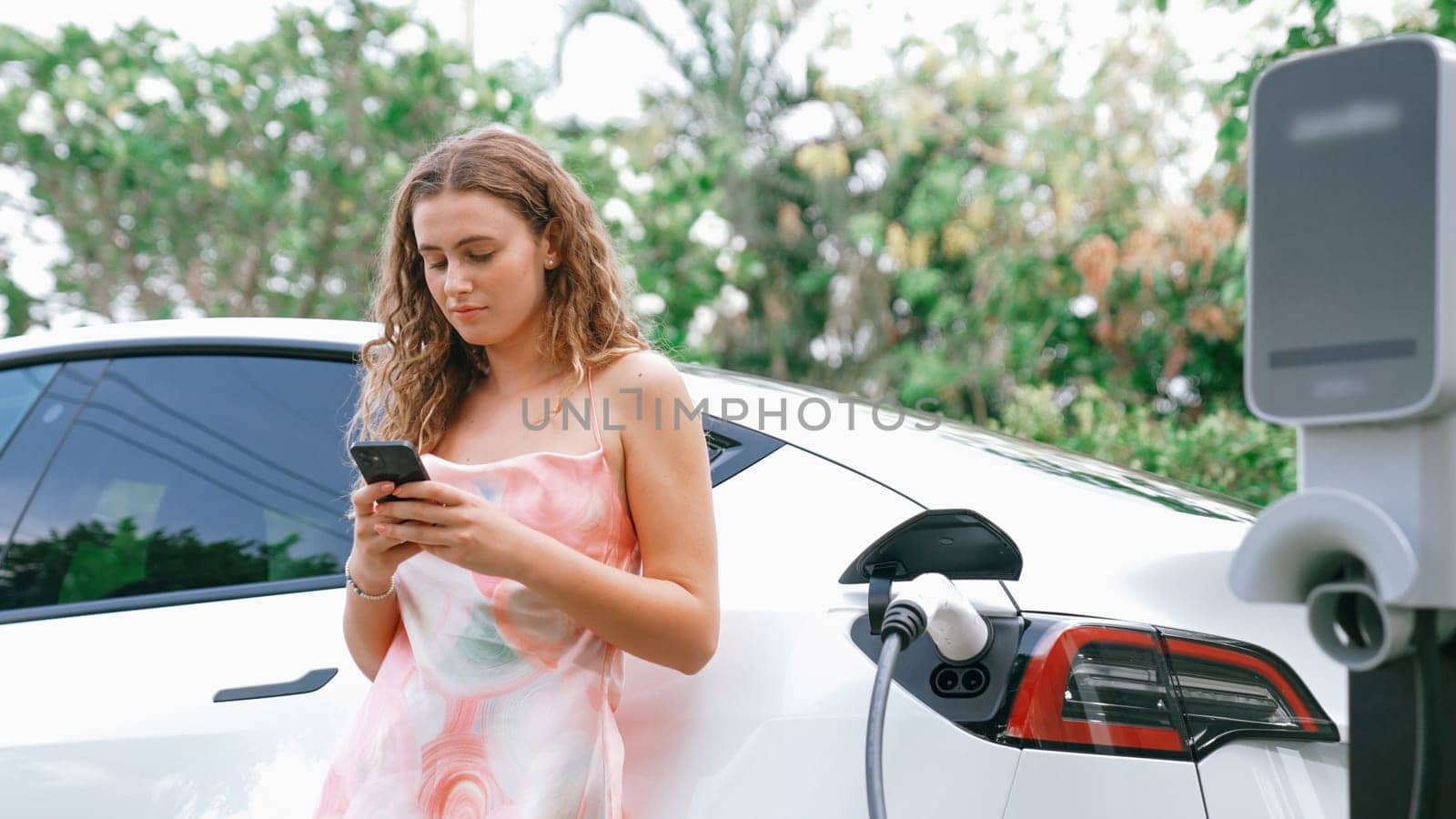 Young woman use smartphone to pay for electricity at public EV car charging station in nature. Modern environmental and sustainable automobile transportation lifestyle with EV vehicle. Synchronos