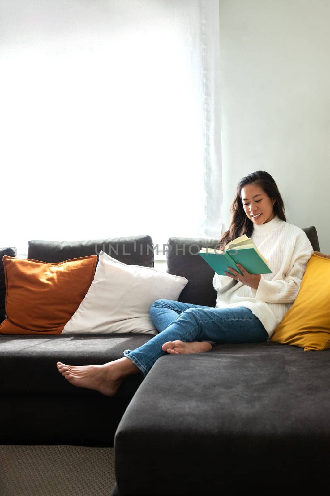Portrait of happy, smiling Asian woman relaxing at home reading a book sitting on the sofa. Vertical image. Copy space. by Hoverstock