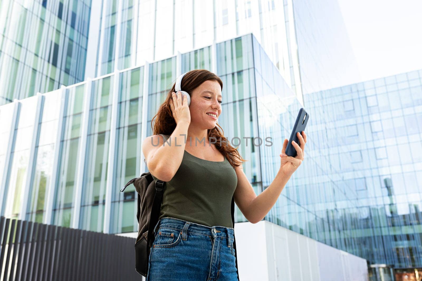 Redhead female university student with backpack listening to music using phone app and headphones standing outdoors in college campus. Copy space. Education and technology concepts.
