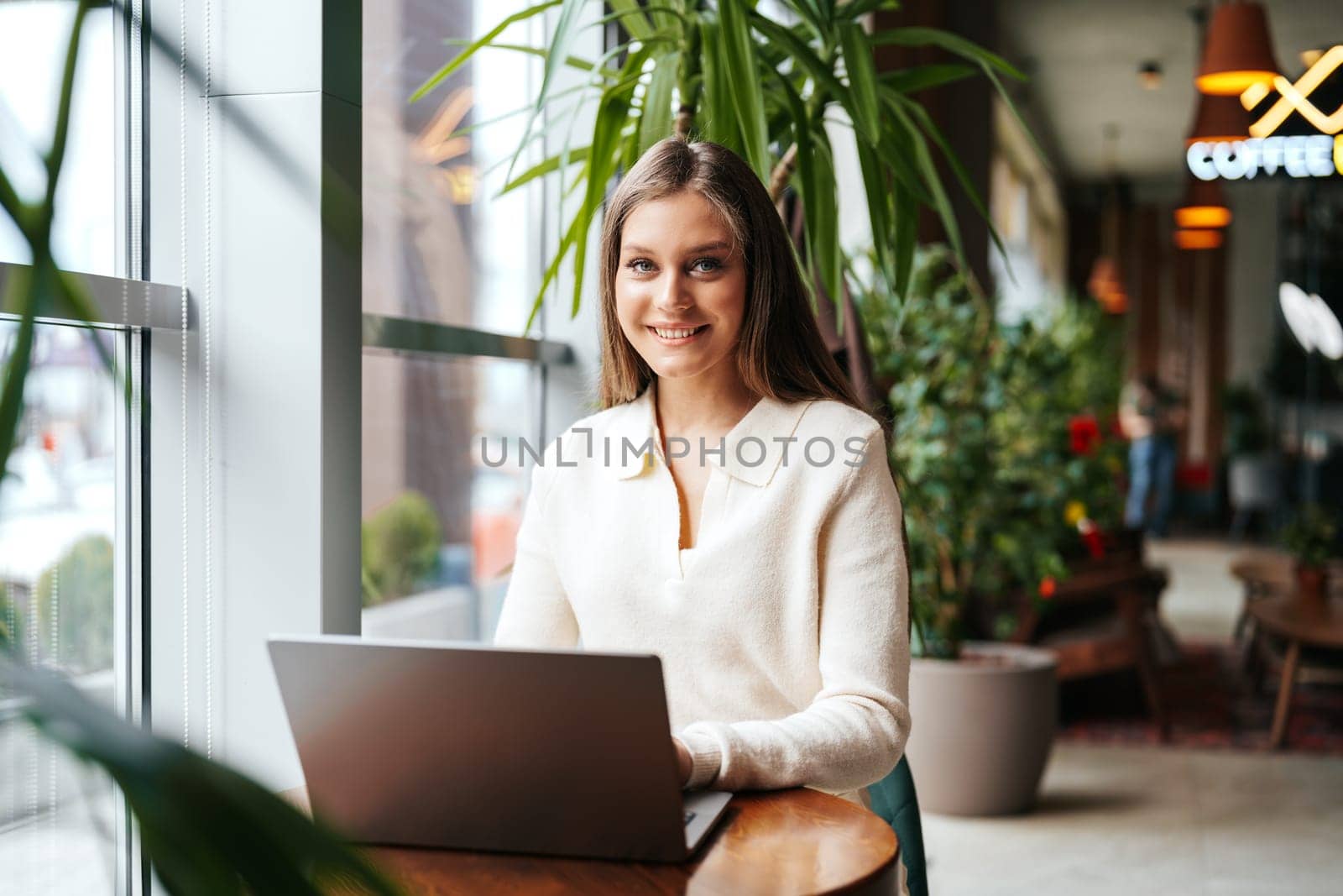 A cheerful young woman is seated at a wooden table in a well-lit cafe, working on her laptop. Her attention is momentarily diverted away from the screen as she smiles towards someone or something, giving off a pleasant and relaxed vibe amidst the indoor greenery that adorns the environment.