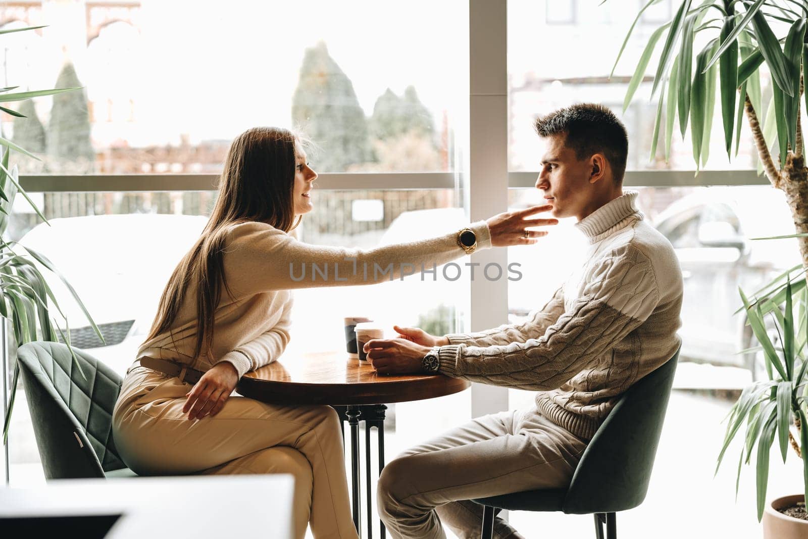 A man and a woman are seated across from each other at a wooden table in a well lit cafe with large windows and indoor plants. They are engaged in a relaxed conversation while sipping from coffee cups, suggesting a close and comfortable relationship between them.