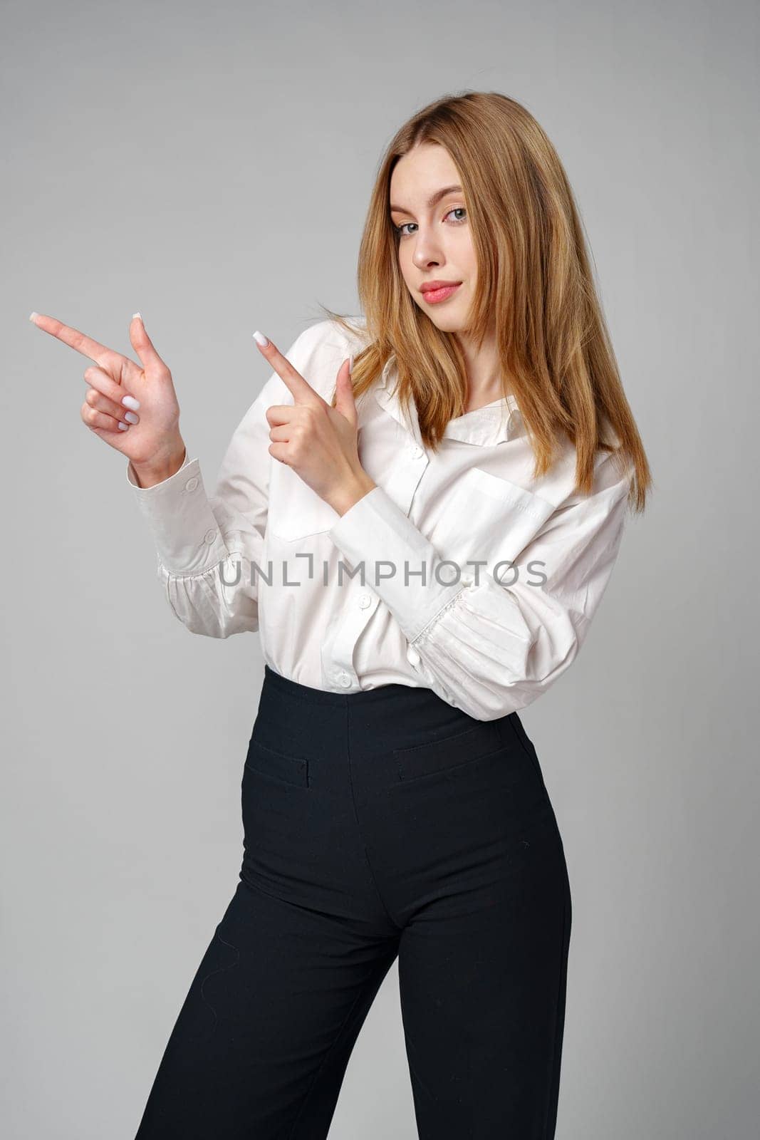 Young blonde woman in formal outfit pointing to the side against gray background in studio