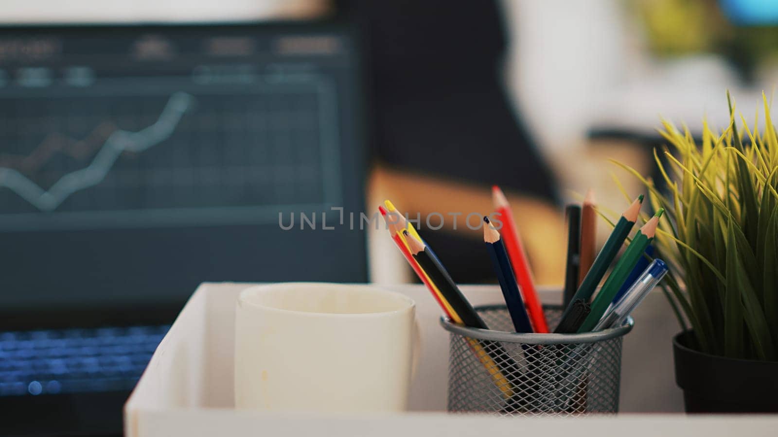 Focus on desk belongings in modern office with notebook in blurry background showing business diagram and figures, close up. Pencil holder, mini house plant, empty coffee mug and graphs on laptop