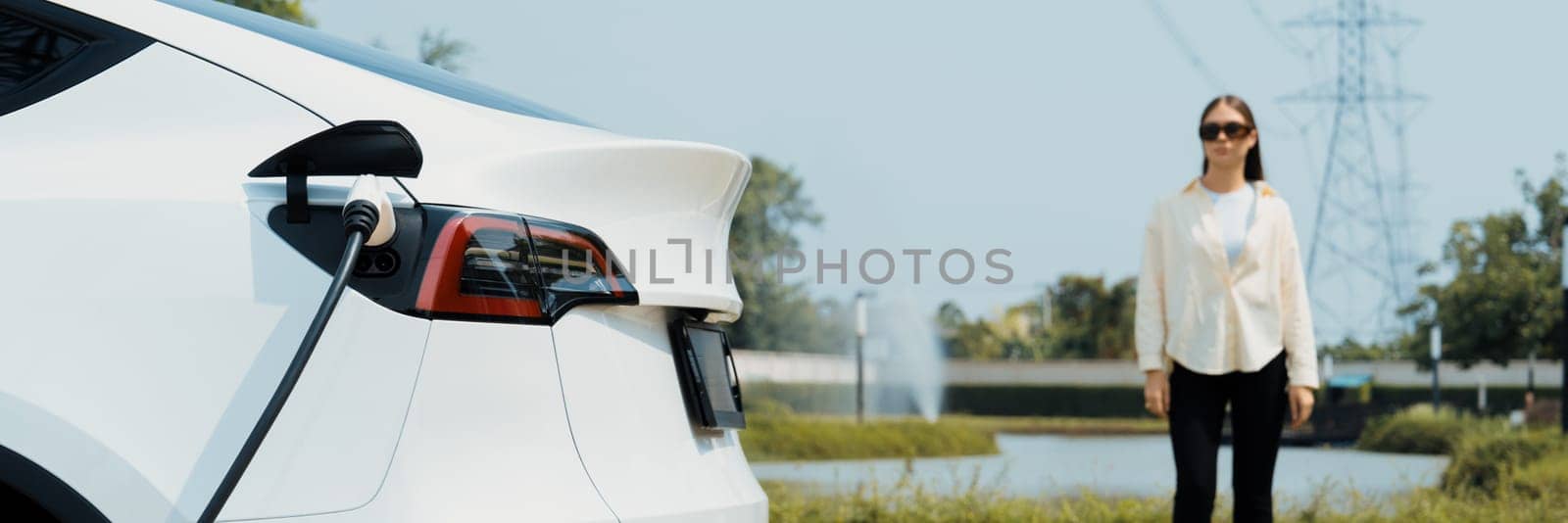 Young woman recharge EV car battery at charging station connected to power grid tower electrical industrial facility as electrical industry for eco friendly vehicle utilization. Panorama Expedient