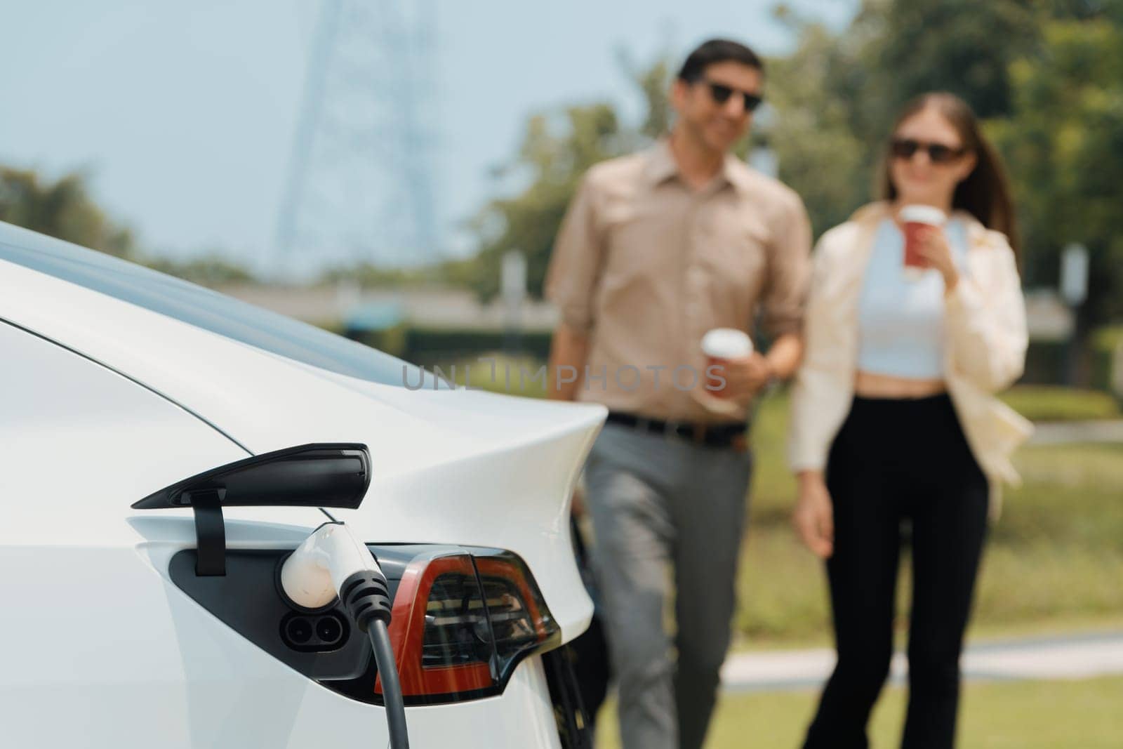 Young couple with coffee cup, recharge electric car's battery from EV charging station in green city park. Sustainable and eco friendly EV car with urban and shopping lifestyle. Expedient