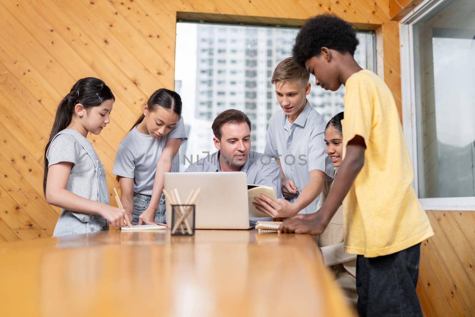 Teacher sitting and giving a lecture on lesson to children in classroom using laptop, talking to children about content in notebook. Students asking about subject instructor teach them. Edification.