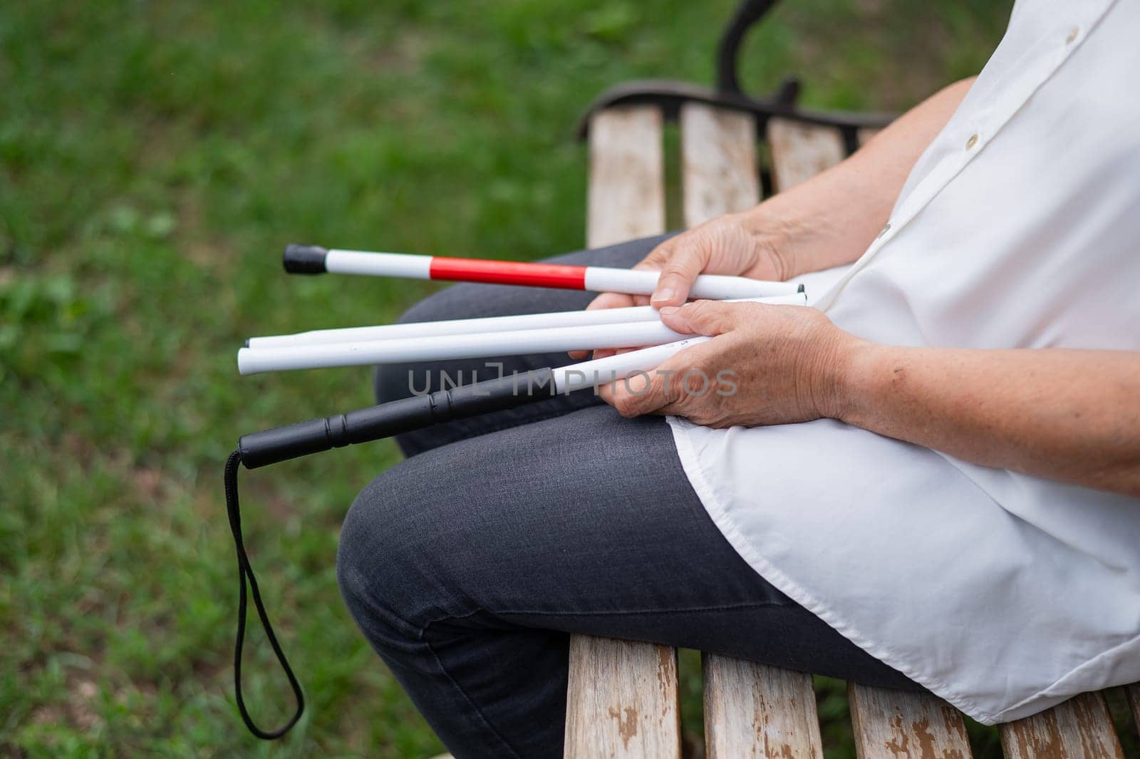 An elderly blind woman sits on a bench in the park. Close-up of female hands with folded tactile cane
