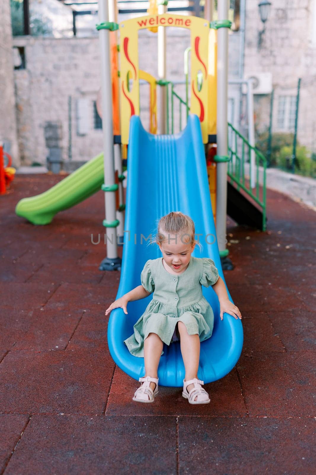 Little smiling girl sitting at the bottom of a plastic slide on a playground in the yard. High quality photo