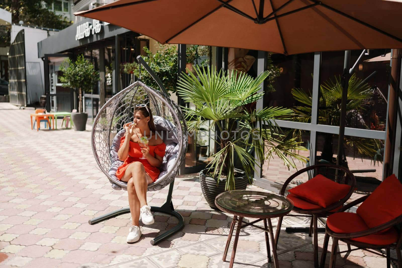 A woman is sitting in a hammock on a patio. The patio is surrounded by potted plants and has a few chairs and tables. The woman is drinking a beverage and she is enjoying her time