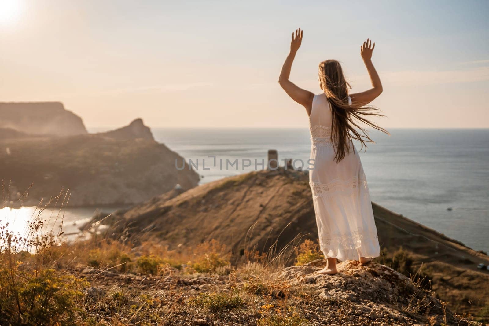 A woman in a white dress stands on a rocky hill overlooking the ocean. She is smiling and she is happy