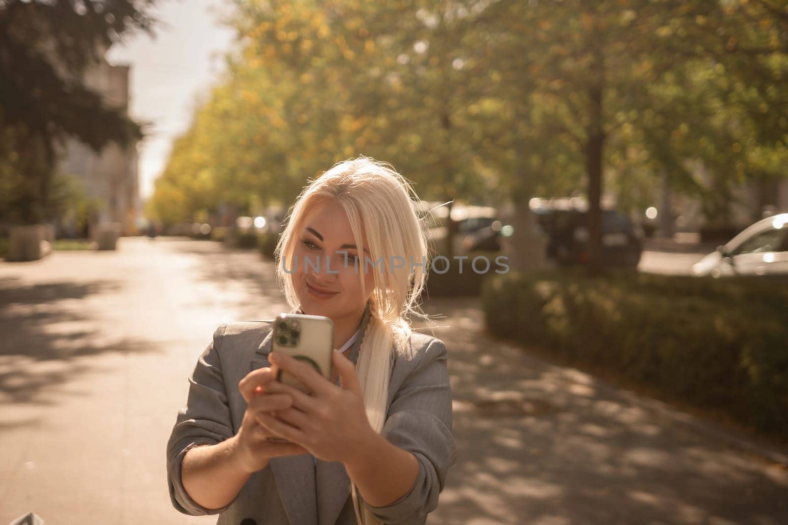 A woman is taking a picture of herself with her cell phone. She is wearing a gray jacket and scarf. The scene is set in a city with trees and cars in the background