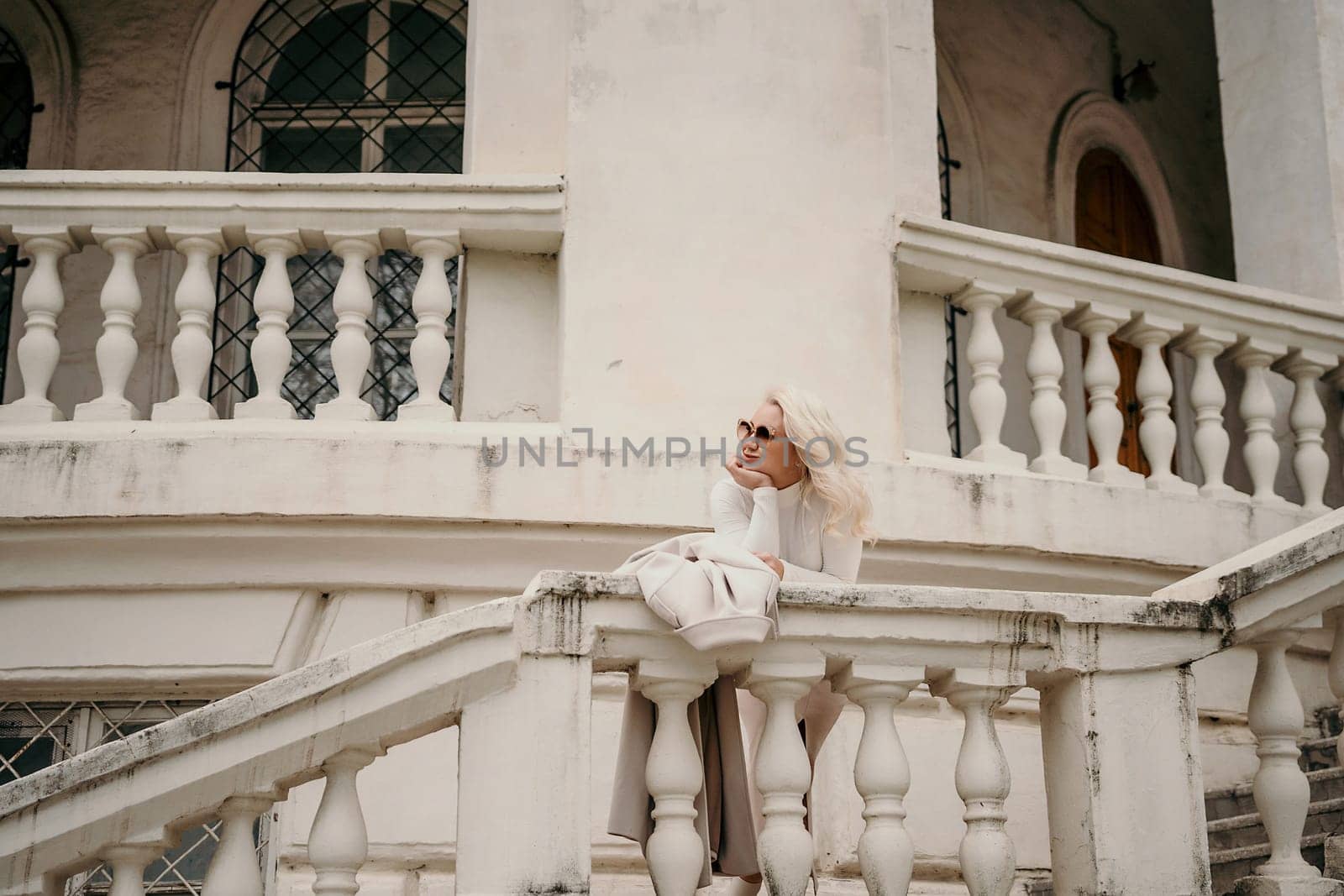 A woman is sitting on a white railing, looking out at the street. She is wearing sunglasses and a white shirt. The scene is set in front of a building with white pillars. by Matiunina