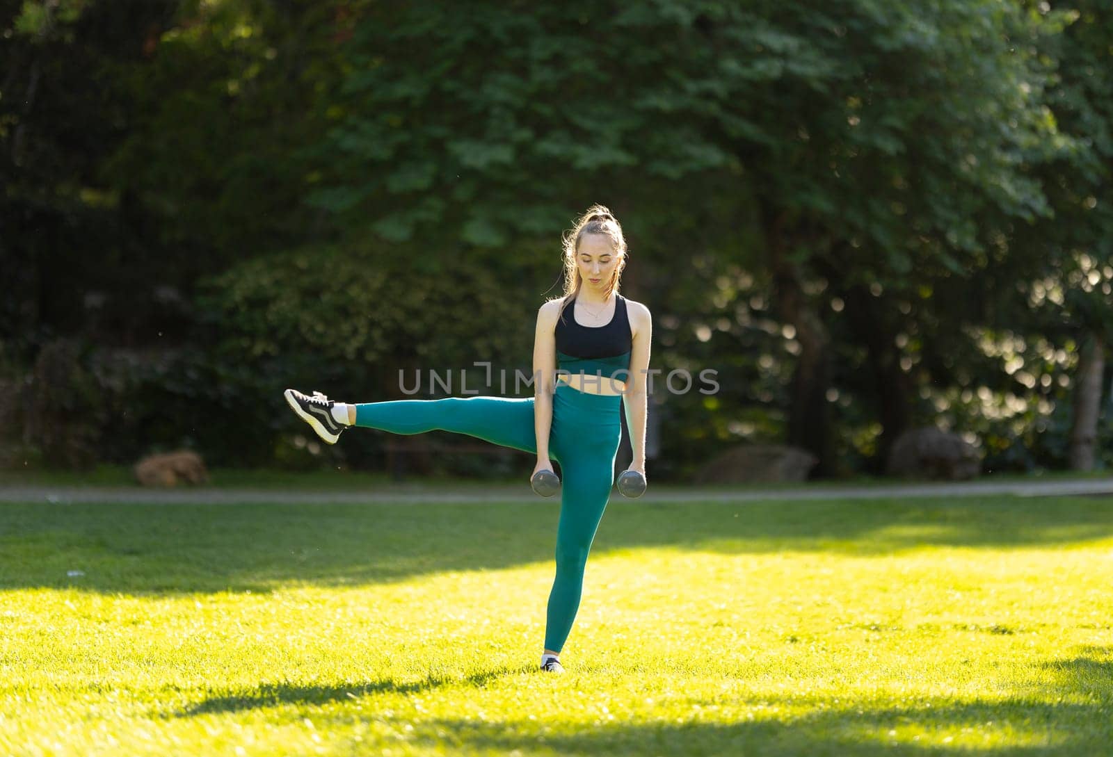 A woman is doing a yoga pose in a park. She is wearing a black tank top and green leggings