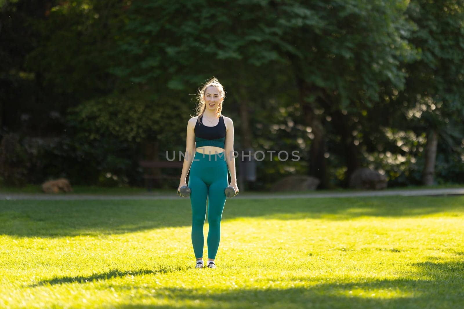 A woman is standing in a park, holding two dumbbells by Studia72