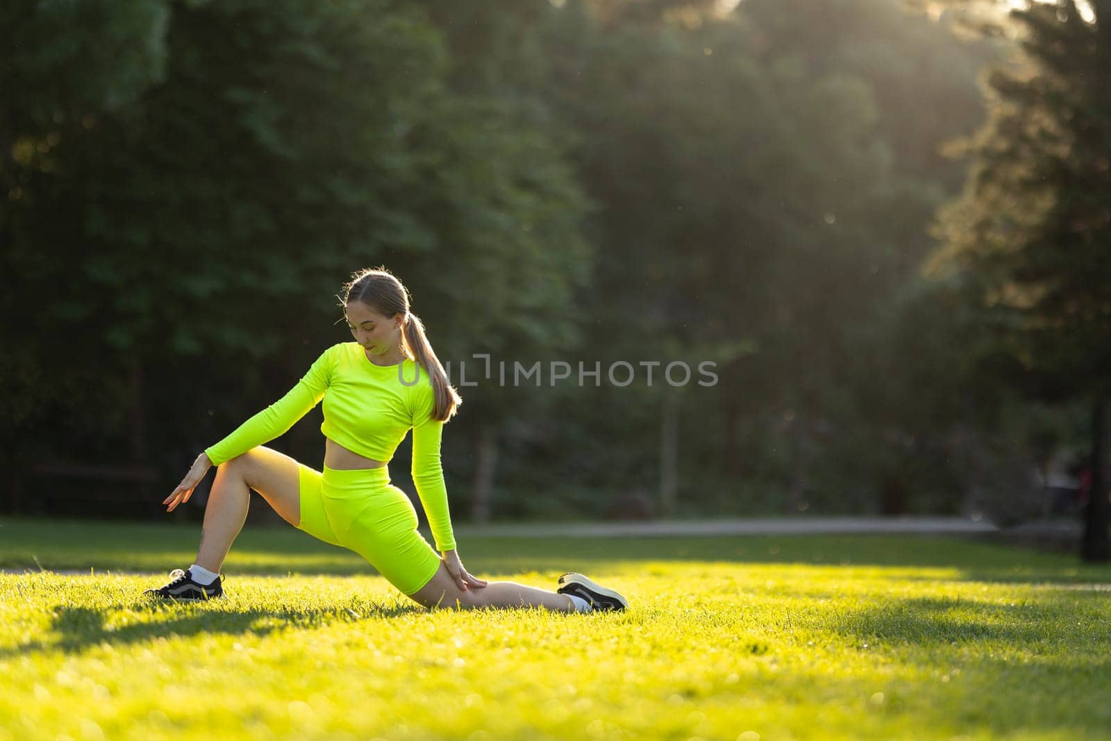 A happy woman in a neon yellow outfit is leisurely stretching her legs on the grass in a park, surrounded by lush greenery and other people enjoying nature