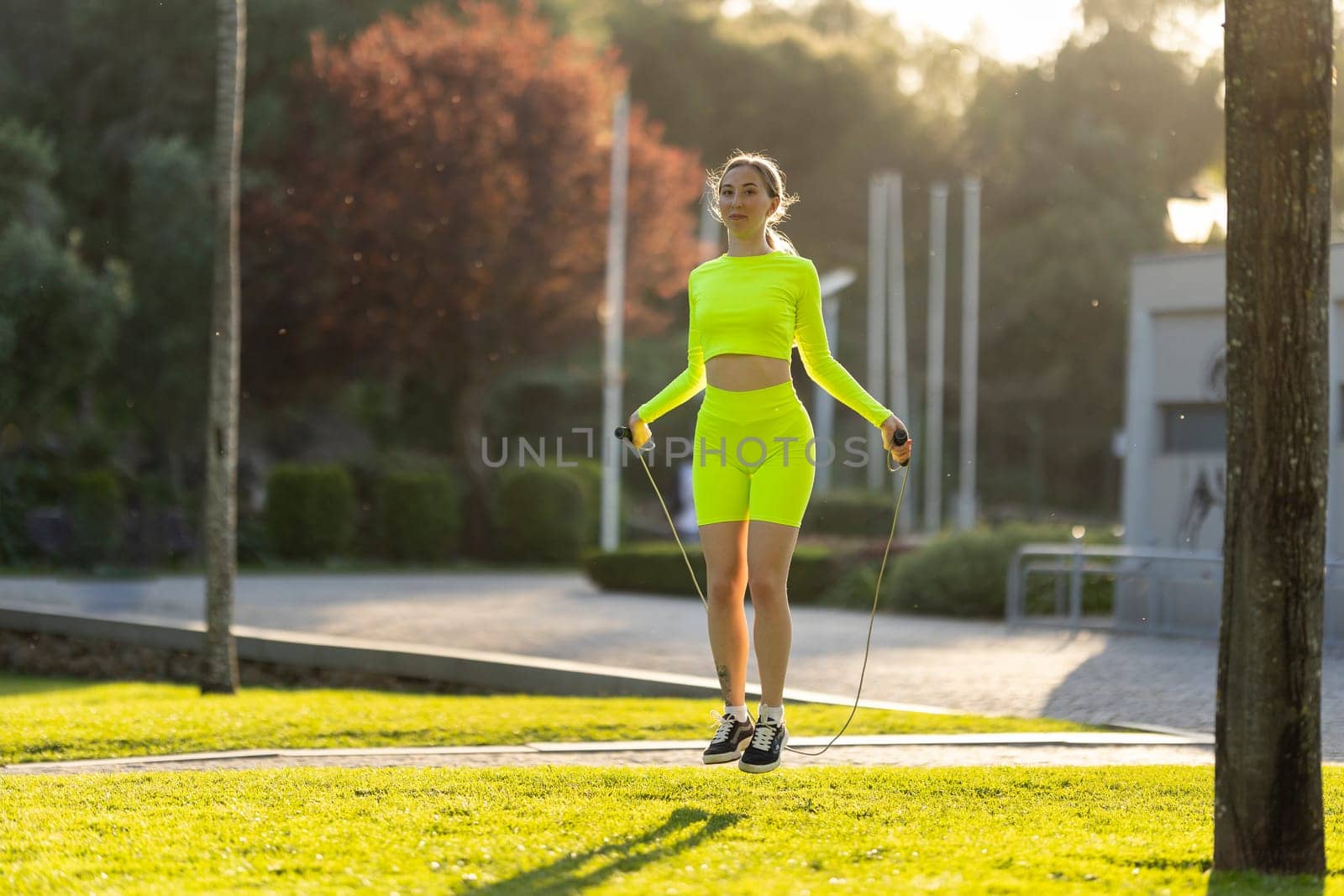 A woman in a neon yellow outfit is jumping rope in a park. The scene is bright and cheerful, with the woman's outfit and the green grass creating a sense of energy and positivity