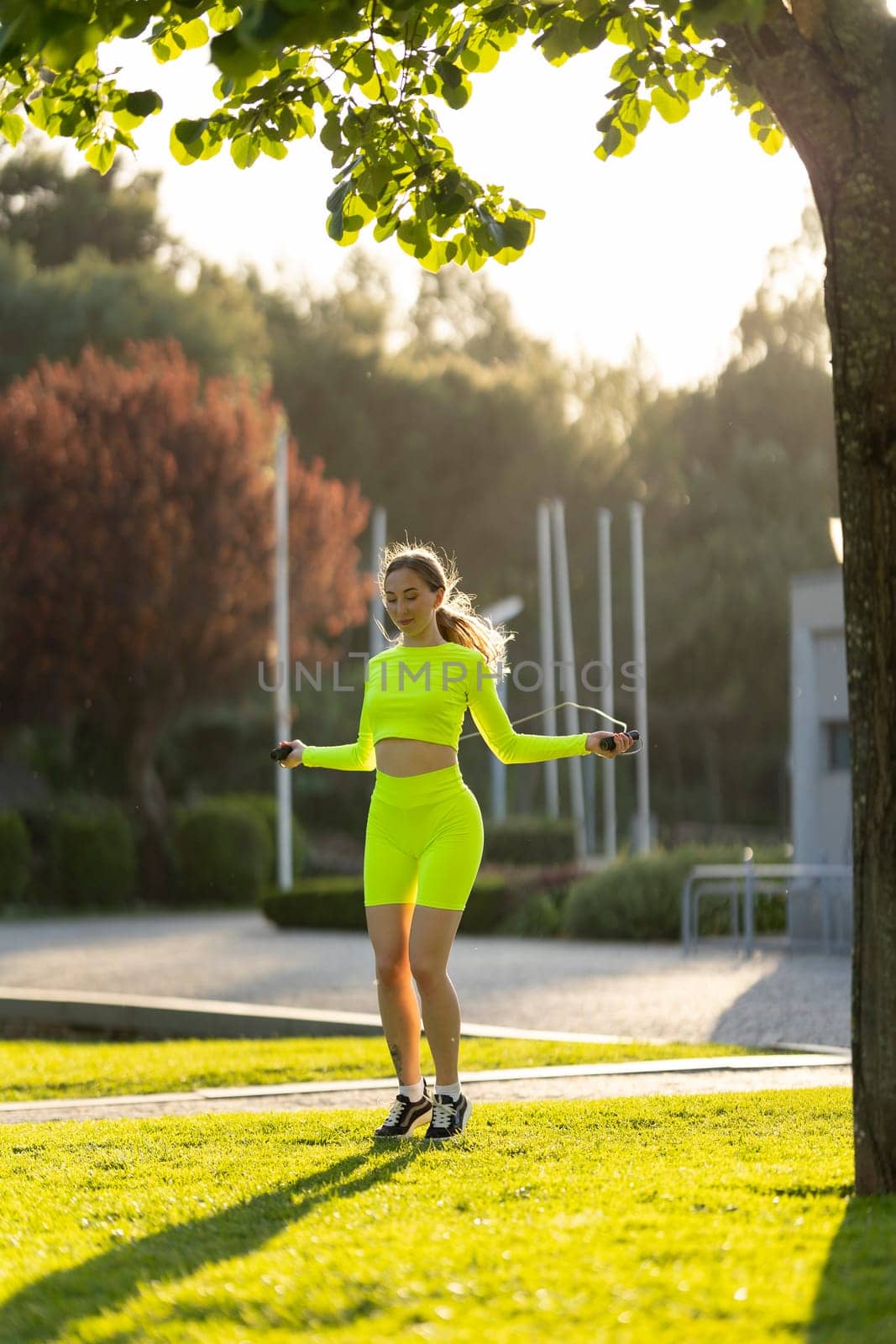 A woman in a neon yellow outfit is jumping rope in a park. The scene is bright and cheerful, with the sun shining down on the woman and the grass