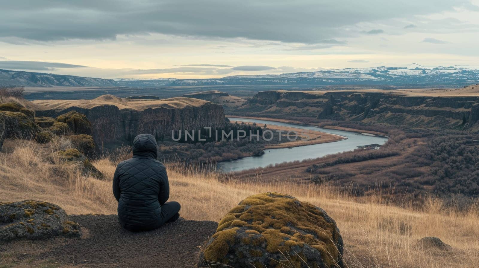 Individual sitting in solitude on a hill, overlooking a tranquil valley with a river snaking through it at dusk
