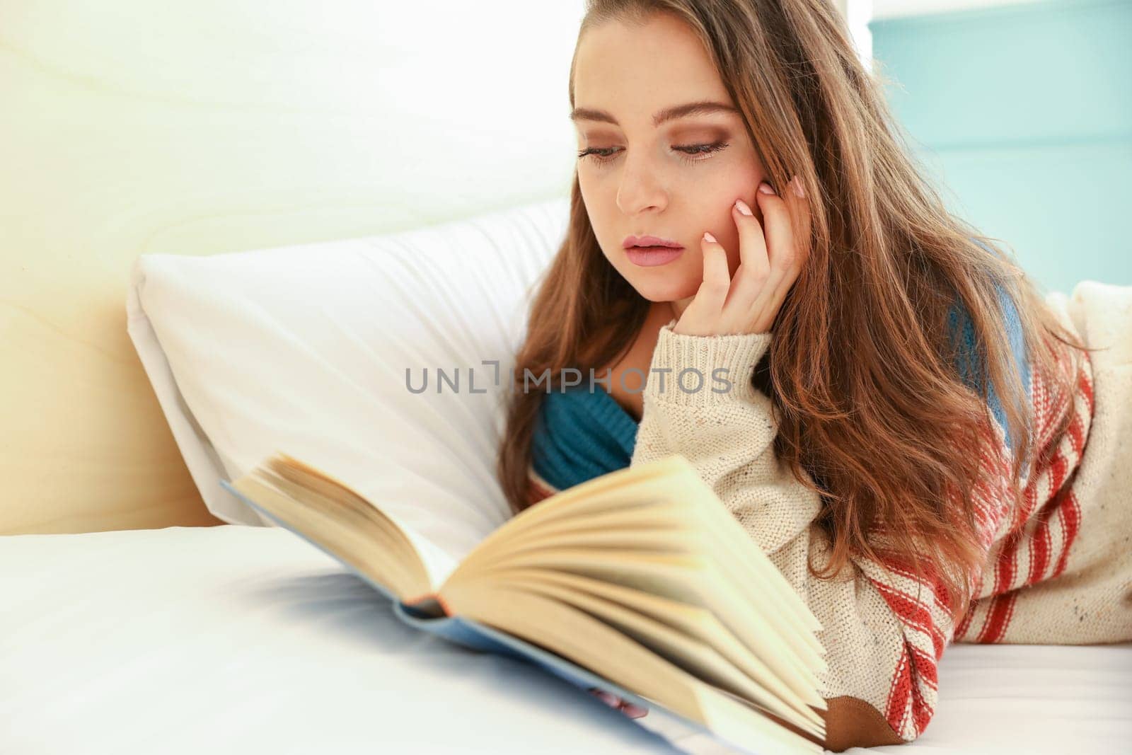 Young model reading a book on the bed, lying face down, dressed in a sweater, resting her head in her hand.