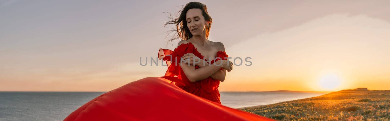 woman red dress standing grassy hillside. The sun is setting in the background, casting a warm glow over the scene. The woman is enjoying the beautiful view and the peaceful atmosphere. by Matiunina