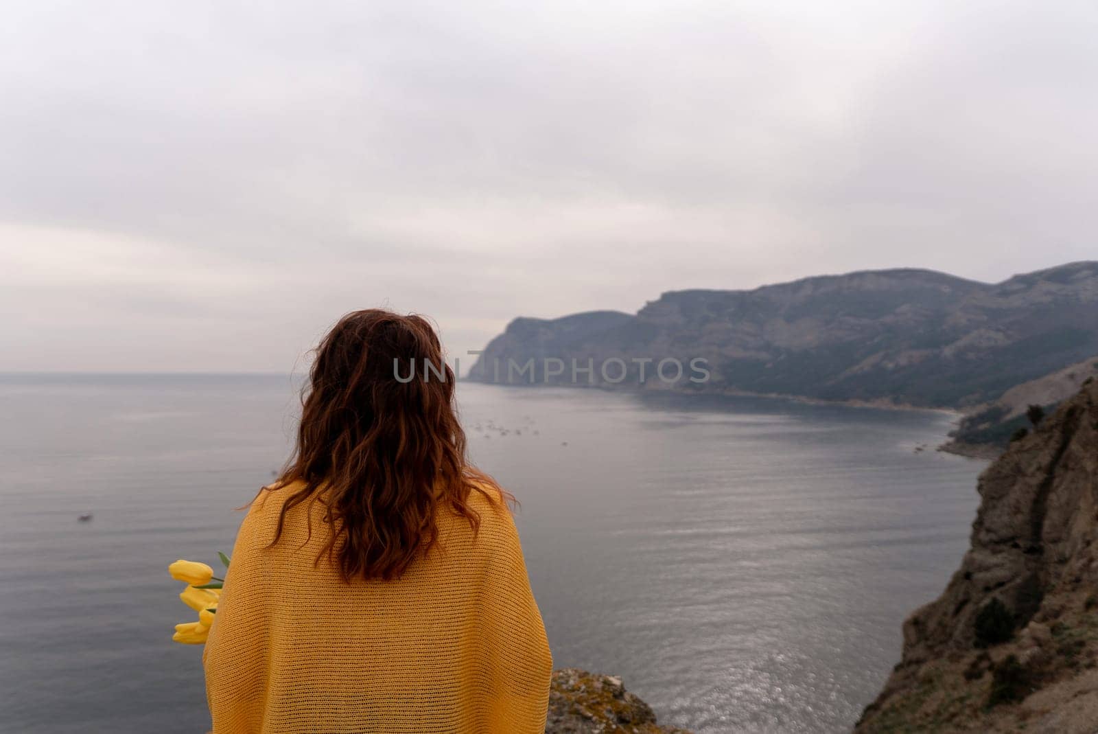 Rear view of a woman with long hair against a background of mountains and sea. Holding a bouquet of yellow tulips in her hands, wearing a yellow sweater by Matiunina