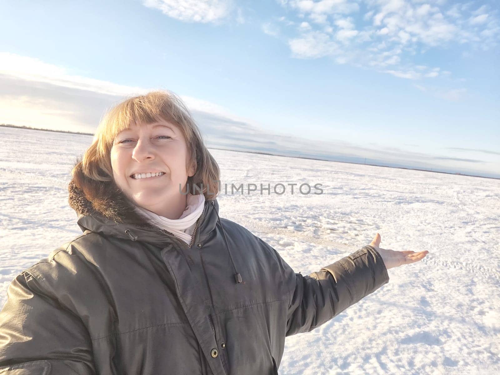 A cheerful middle aged woman in a winter coat taking selfie on nature outdoors in sunny day with blue sky